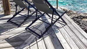Une terrasse en bois avec trois chaises longues donnant sur une mer turquoise, sous un ciel dégagé.