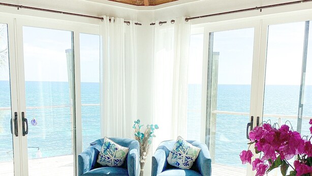 Une pièce avec deux fauteuils bleus, une table ronde blanche et une vue sur la mer, visible à travers de grandes portes vitrées.