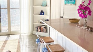 Une cuisine avec un plafond en bois, des meubles blancs, un coin bar avec des tabourets et une vue sur l’océan grâce à de grandes fenêtres.