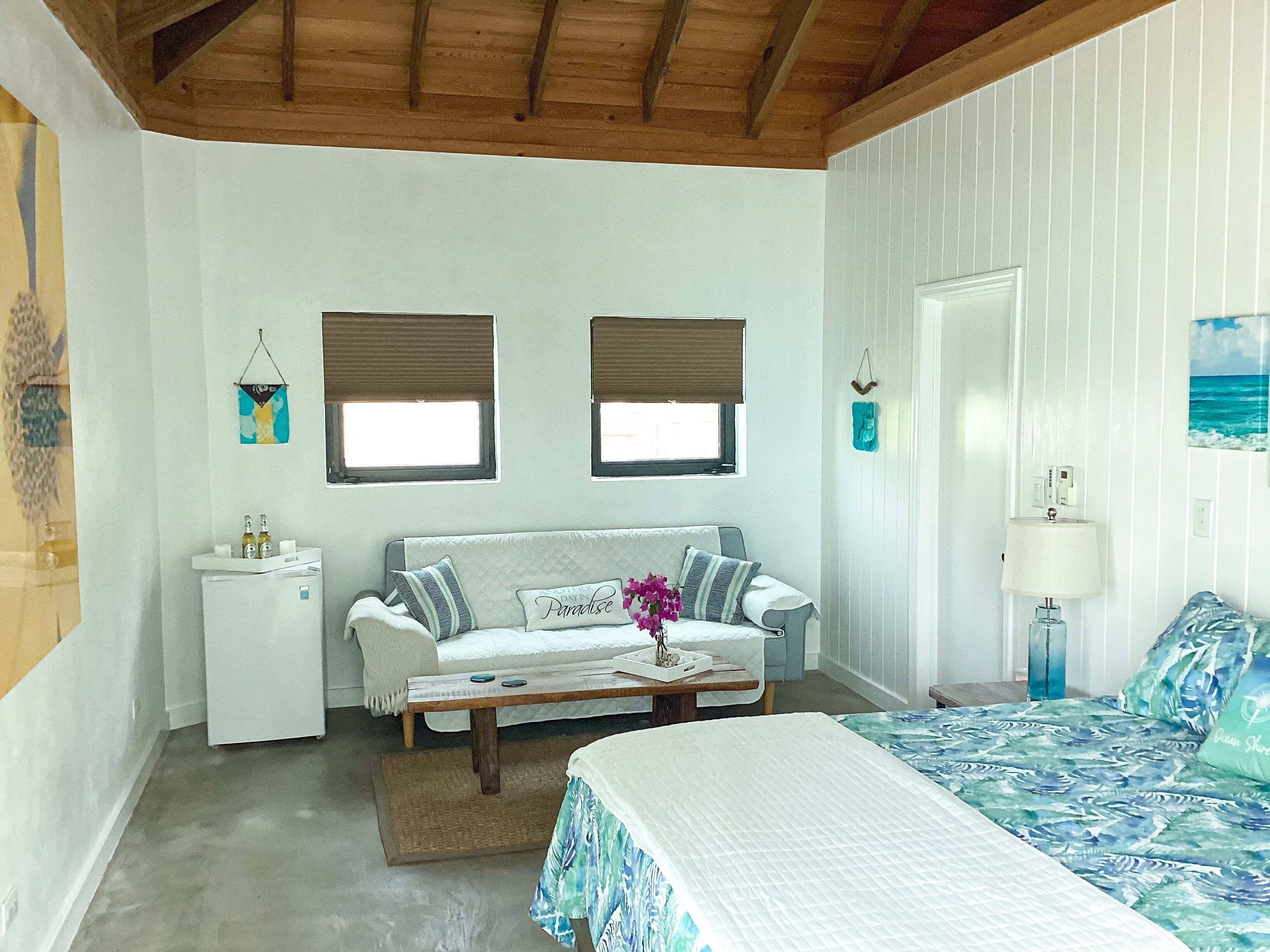 A bedroom with a bed, a sofa, a small table, and a nightstand. There are two windows with blinds, a door, and a wall-mounted lamp.
