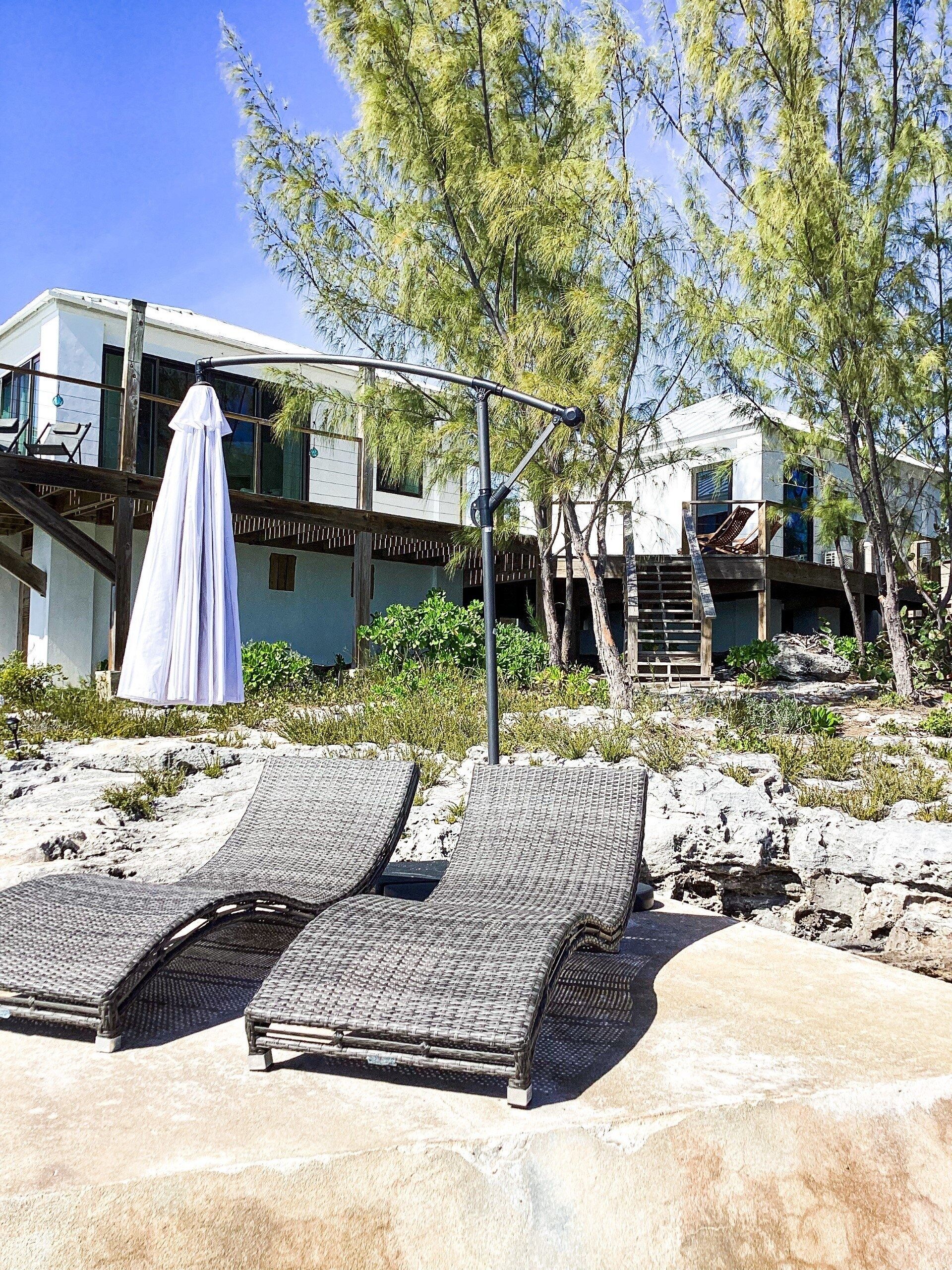 Une propriété située en bord de mer, comprenant deux chaises longues, un parasol et un bâtiment moderne doté d’un balcon.