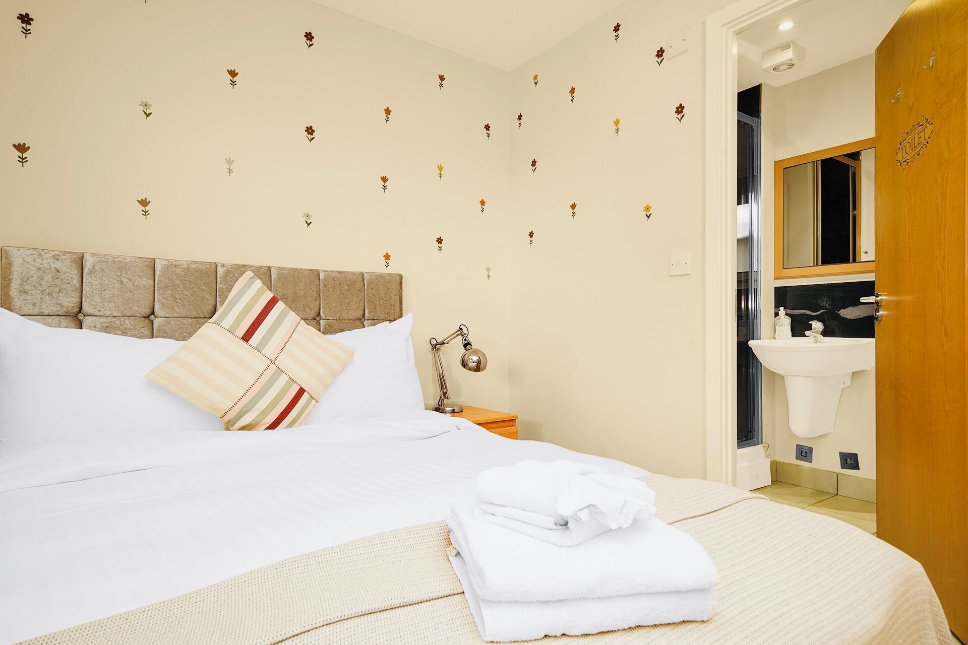 A hotel room with a bed, a nightstand, a lamp, a bathroom with a sink, and a door labeled "TOILET".