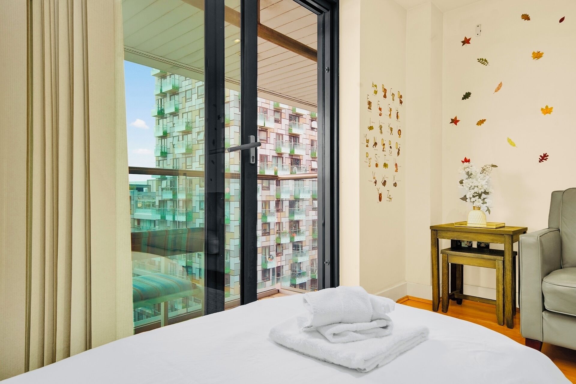 Une chambre d’hôtel avec un grand lit, une vue sur un immeuble à travers une porte vitrée, une petite table en bois et un canapé gris.