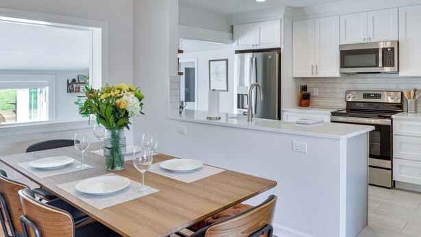 Une cuisine moderne avec des meubles blancs, des appareils en acier inoxydable et une table à manger en bois pour quatre personnes.