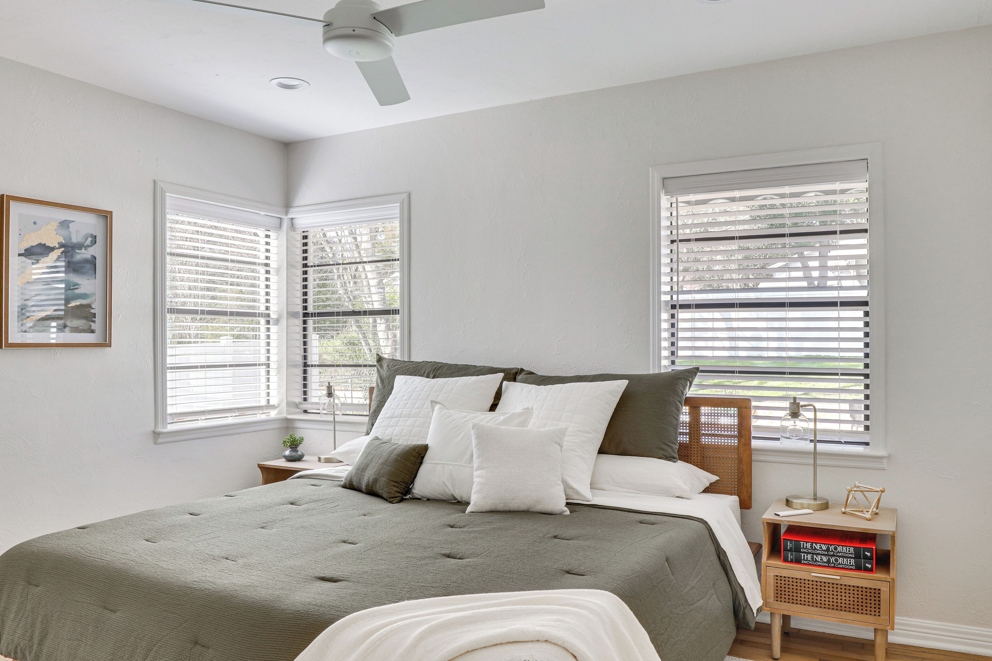 A bedroom with a bed, two windows with blinds, a bedside table with a lamp, and a framed picture on the wall.