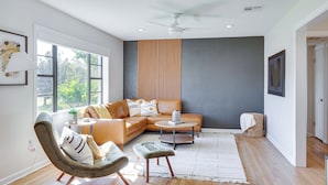 Un salon moderne avec un canapé d’angle, une table basse ronde et un fauteuil. On y trouve un ventilateur de plafond, une fenêtre donnant sur un espace verdoyant et une étagère fixée au mur.