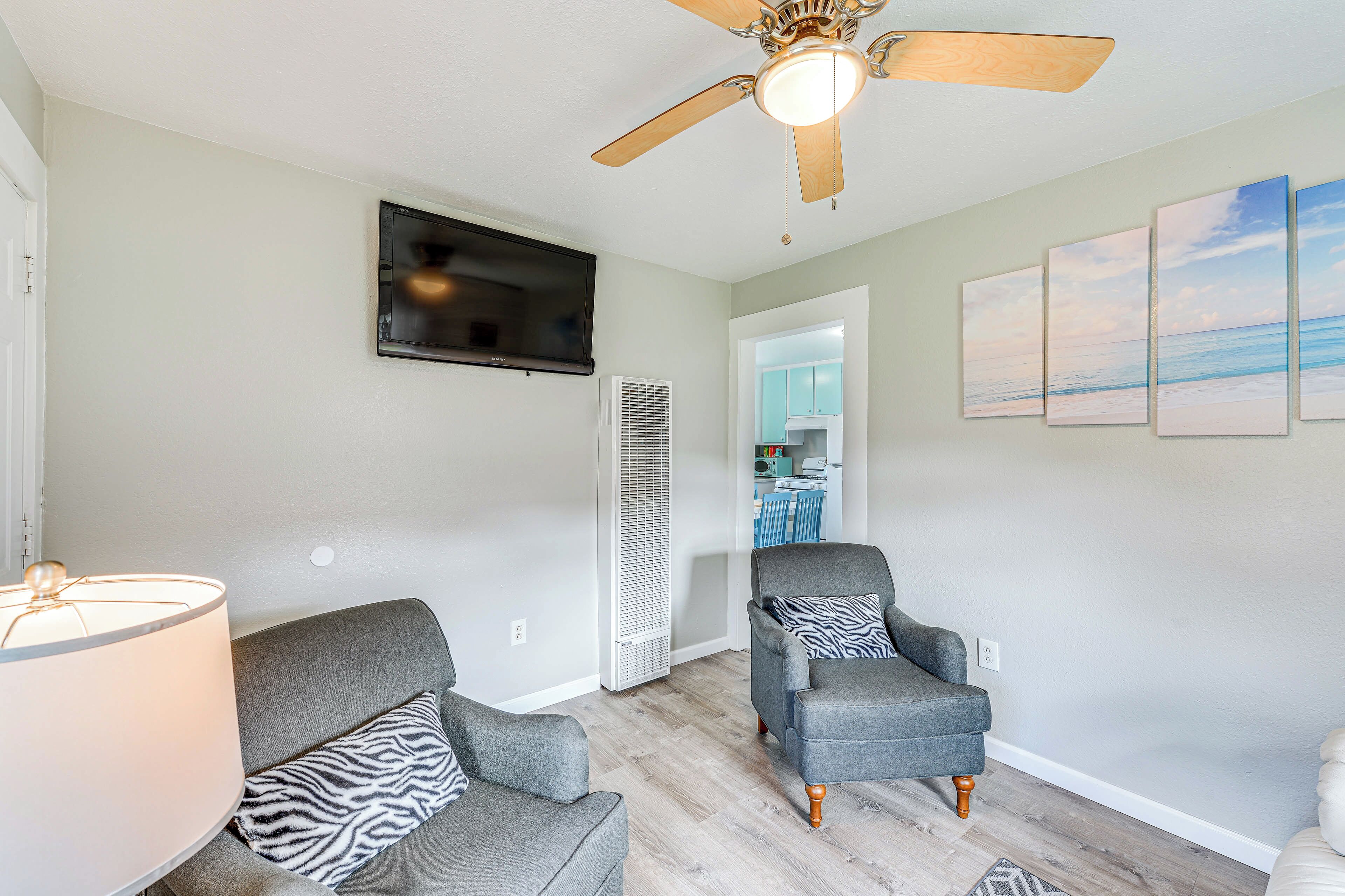 A living room with two grey armchairs, a ceiling fan, a TV mounted on the wall, and a view into the kitchen.