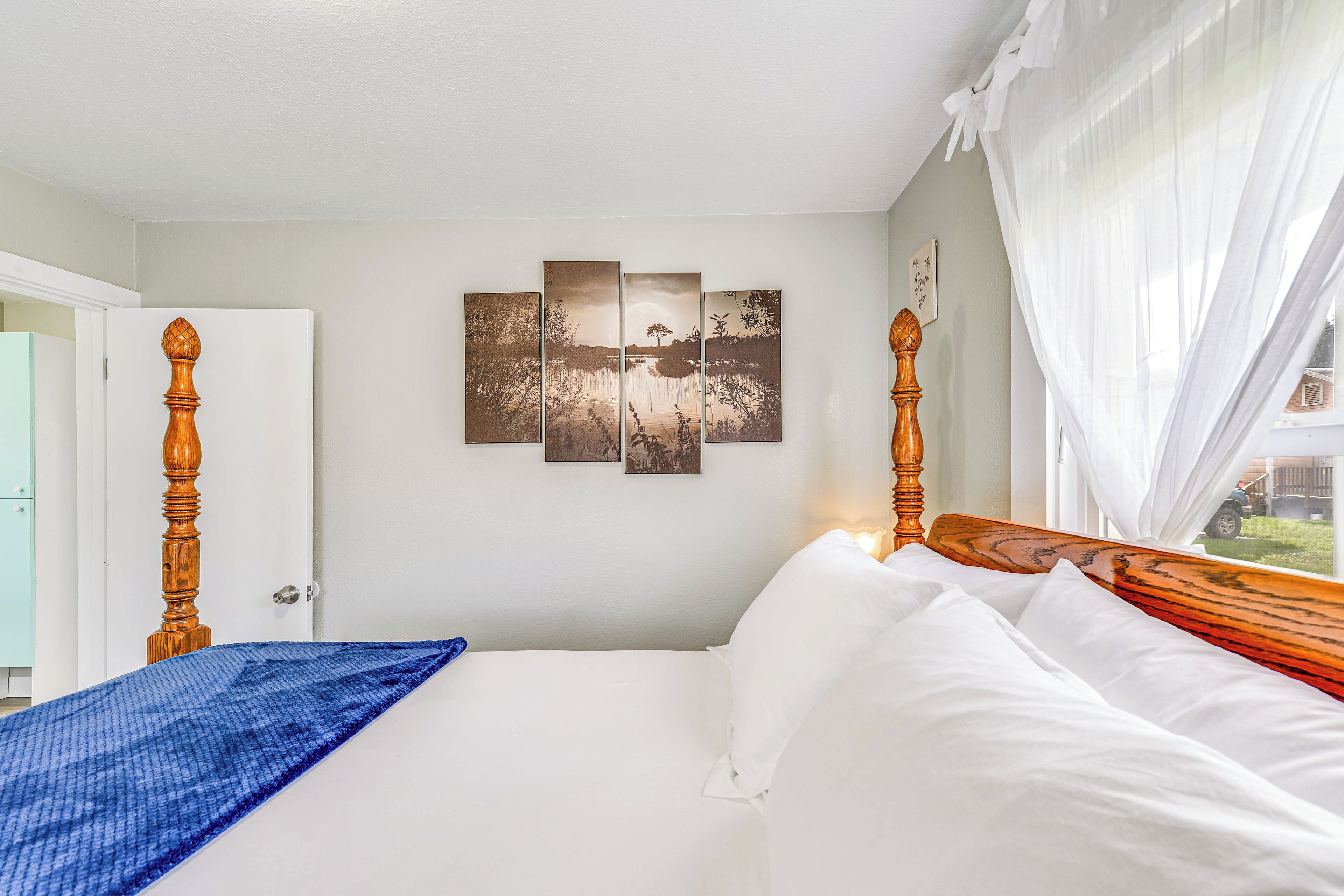 A bedroom with a bed, a wooden headboard, a window with sheer curtains, and a wall-mounted artwork.