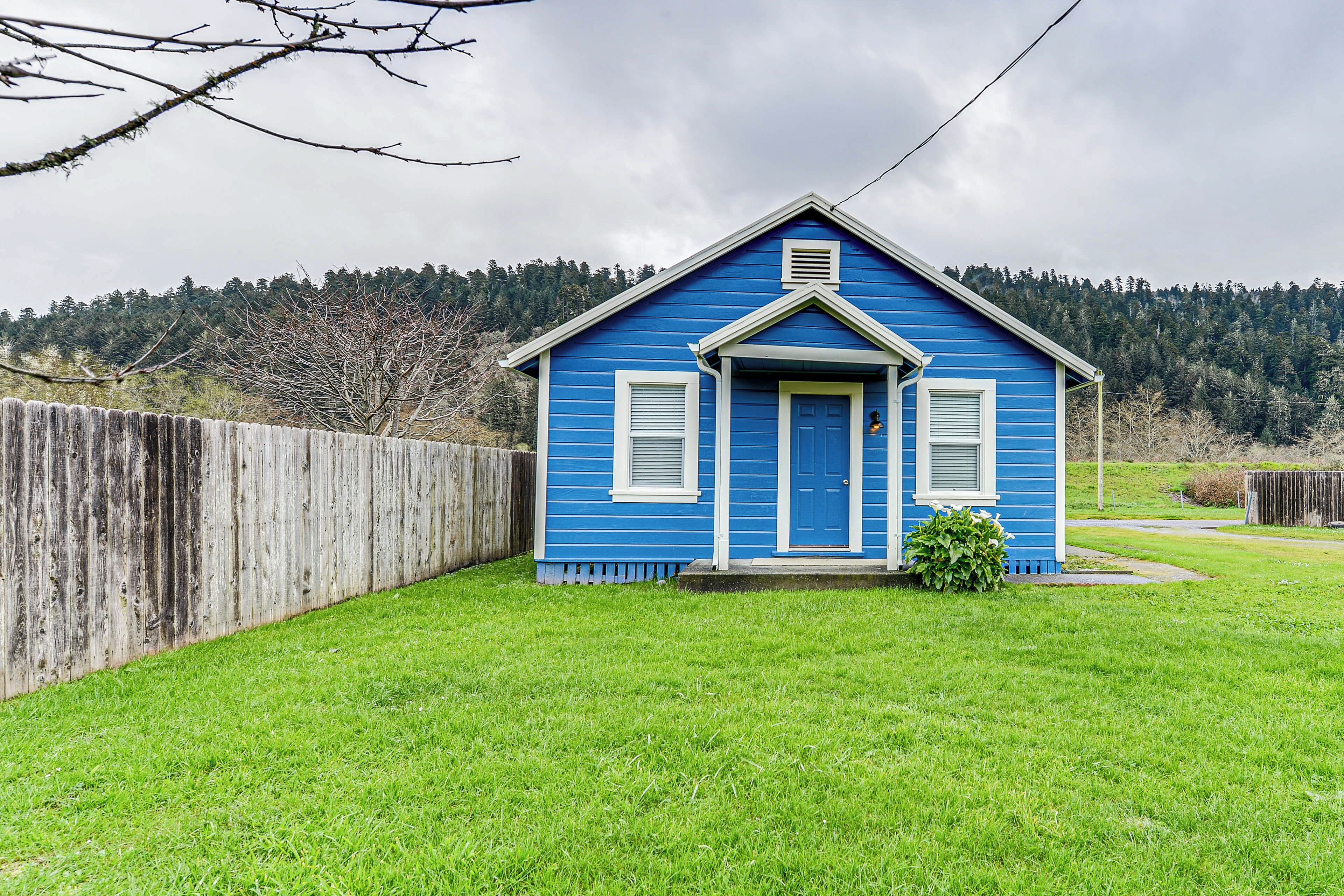 Una pequeña casa azul con puerta principal y dos ventanas, rodeada por una cerca de madera y un jardín cubierto de césped.