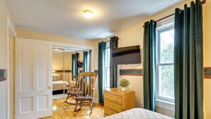 Un dormitorio con cama, sillón mecedora, cómoda, televisor montado en la pared y ventana con cortinas.