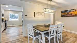 A dining area with a wooden table and white chairs, a pendant light, a wall-mounted sign, and a painting. - Riverfront Michigan Cottage w/ Fire Pit & Kayaks! (Beaverton)
