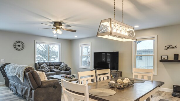 A spacious living room with a dark brown leather sofa, a white dining table with chairs, a ceiling fan, and a large flat-screen TV. - Riverfront Michigan Cottage w/ Fire Pit & Kayaks! (Beaverton)