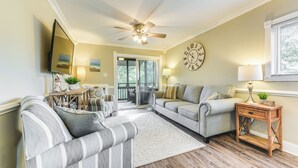 A living room with a sofa, armchair, and a coffee table. There is a clock on the wall and a ceiling fan. - Walk to Ocean: Unit w/ 2 Balconies in Myrtle Beach (Myrtle Beach)