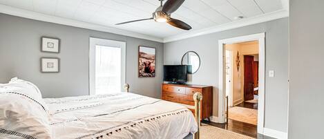 Une chambre à coucher comprenant un lit, un ventilateur de plafond, une commode, un téléviseur et une porte donnant sur une autre pièce.
