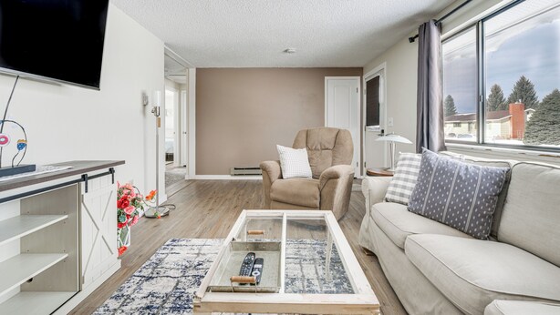 A modern living room with a sofa, a coffee table with a remote control, a TV mounted on the wall, and a view of the outside through a large window. - Dog-friendly Montana Home w/ Deck & Fenced-in Yard (Dillon)