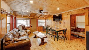 L'intérieur d'un chalet en bois comprend un canapé, une table à manger, des chaises, un ventilateur de plafond et un téléviseur.