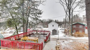 Une terrasse en bois avec des rampes rouges, un petit abri de jardin et une maison rouge, le tout dans un décor enneigé.