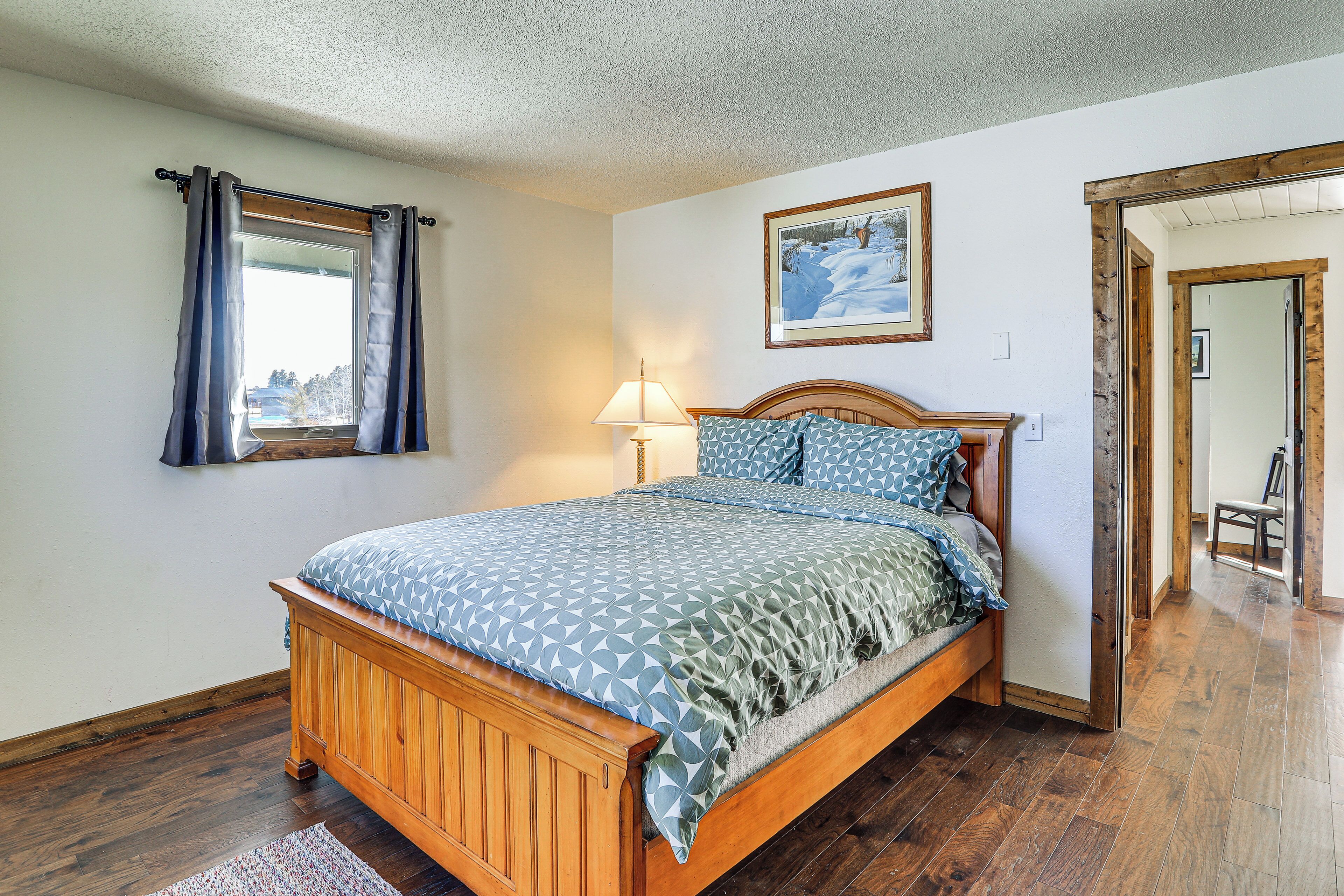 A bedroom with a wooden bed, a window with curtains, a bedside lamp, and a framed picture on the wall.