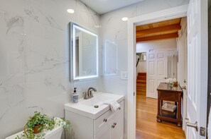 Een badkamer met een witte wastafel, spiegel en marmeren wanden. Er is een houten deur die naar een andere kamer leidt.