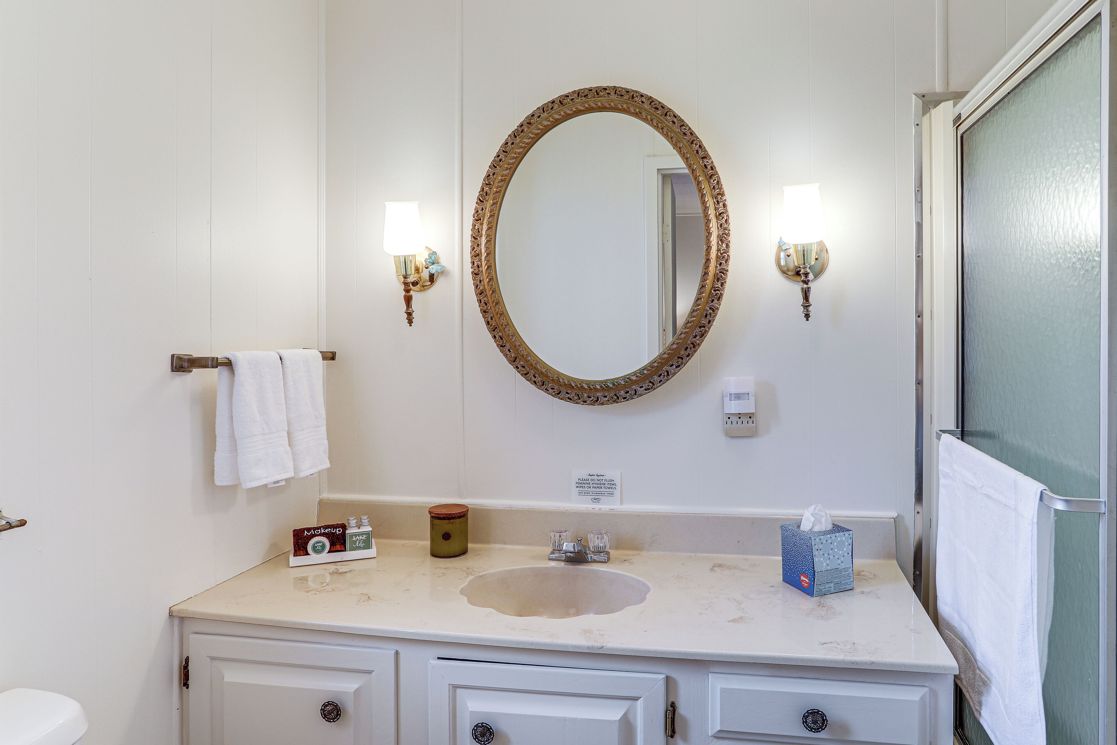Un baño con lavamanos, tocador y un espejo grande con marco decorativo. Hay dos luces fijadas a la pared y un perchero con una toalla blanca.