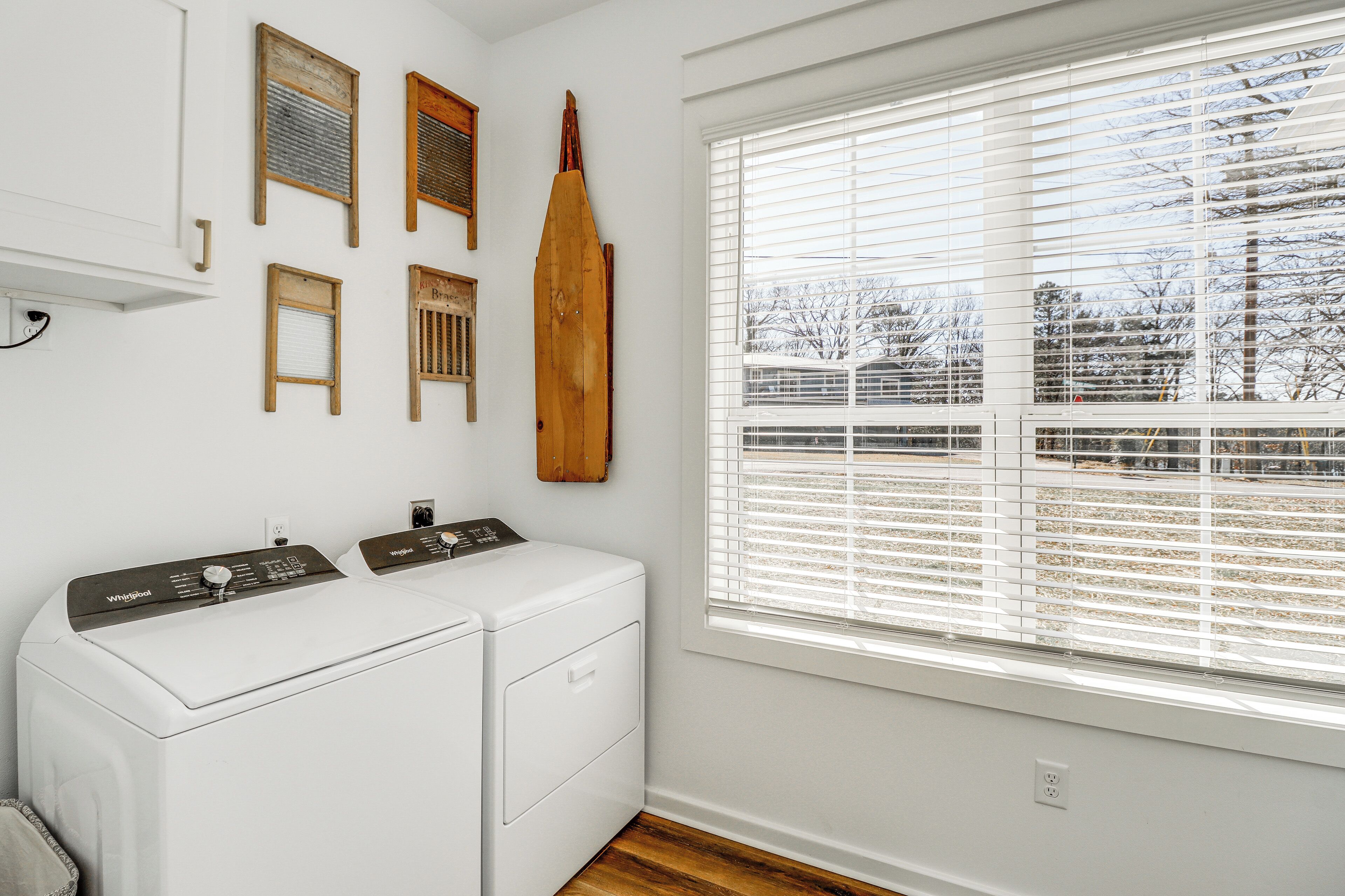 A laundry room with two white Whirlpool washers, wooden wall decorations, and a large window with blinds.