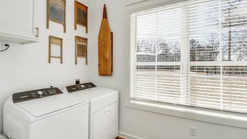 A laundry room with two white Whirlpool washers, wooden wall decorations, and a large window with blinds.