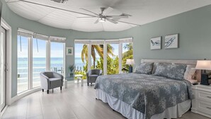 Une chambre avec un grand lit, deux fauteuils, un ventilateur de plafond et une vue sur l’océan.