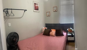 A bedroom with a bed, a fan, a nightstand with a vase, and a window with blinds.