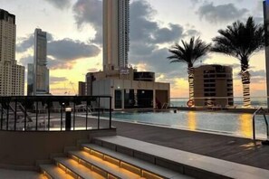Un área de piscina en la azotea con escalones que bajan a la piscina, rodeada de palmeras y con vistas a un horizonte urbano al atardecer.