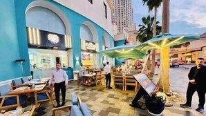 Ein Sitzbereich im Freien mit Tischen und Stühlen, ein kleiner Kiosk und im Hintergrund ein Gebäude mit Rundbogenfenstern.