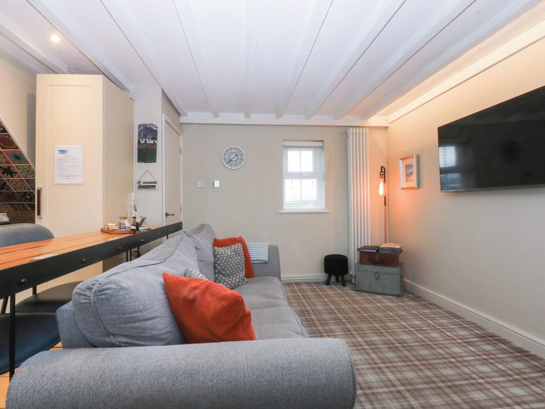 A cozy living room with a grey sofa, a wooden table, a clock on the wall, and a television.
