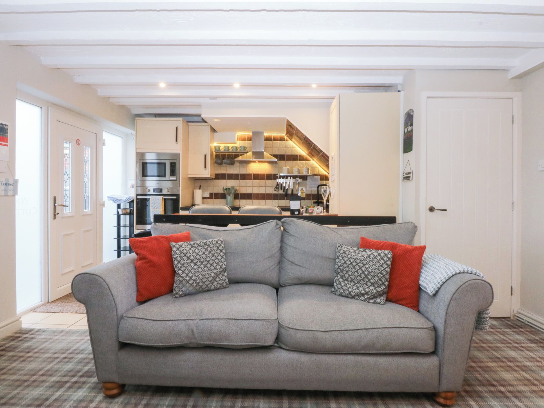 A living room with a grey sofa, red and patterned cushions, and a checkered floor. A kitchen is visible in the background with modern appliances and a tiled backsplash.