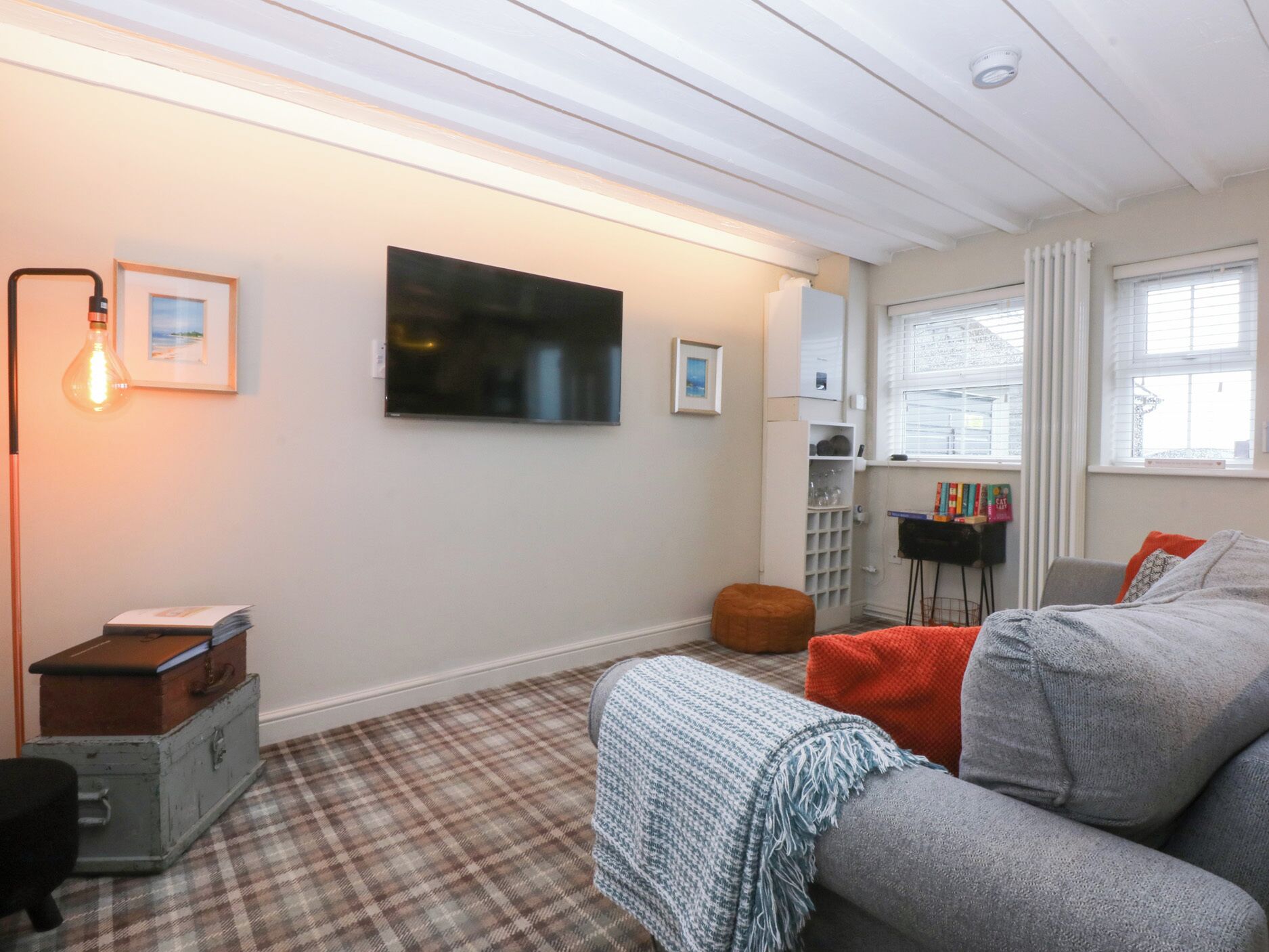 Una habitación acogedora con alfombra escocesa, un sofá gris, un pequeño reposapiés, una estantería y un televisor de pantalla plana montado en la pared.