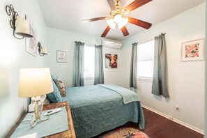 A bedroom with a bed, a ceiling fan, a window with curtains, a wall-mounted air conditioner, and a framed picture.