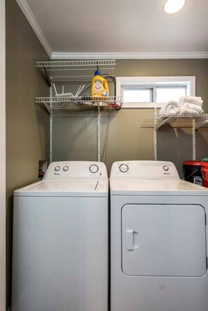A laundry area with two front-loading washing machines, a shelf with a bottle of detergent, and a window above the machines. - Songbird Haven Cozy Country Getaway (Waco)