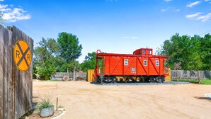 A red caboose with 'ROCK ISLAND' written on it, parked on a gravel area with a wooden fence and a railroad crossing sign nearby. - Chic Caboose in Wimberley Square (Wimberley)