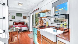 A compact kitchen with a sink, stove, and microwave. A bed with red bedding is visible in the background. - Chic Caboose in Wimberley Square (Wimberley)
