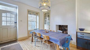 Un coin repas avec une table dressée pour six personnes, des chaises en bois, une nappe à motifs, un buffet bleu et une cheminée en pierre.