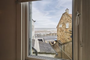 Blick aus einem Fenster auf eine Küstenstraße mit Steinhäusern und einem Strand in der Ferne.