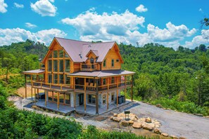 A large wooden house with multiple levels, large windows, and a balcony, surrounded by lush greenery and a clear sky.