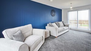 Un salon avec deux canapés blancs, un mur d’accent bleu, une horloge et une grande fenêtre avec des stores.