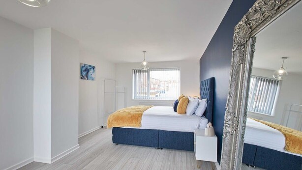 Une chambre moderne avec un grand lit, une table de chevet blanche, un miroir et une fenêtre avec des stores.