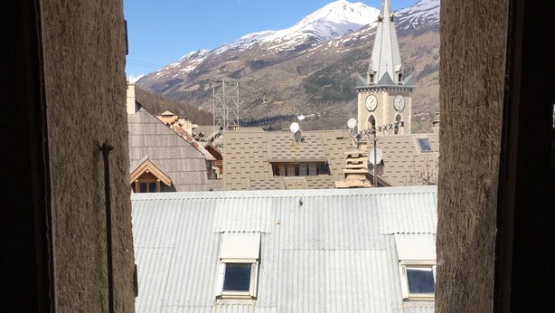 Vista a través de una ventana de un edificio con una torre de reloj, una montaña nevada y un cielo despejado.