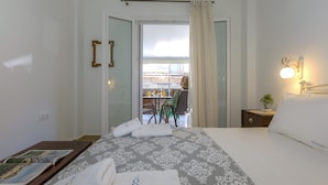 A hotel room with a bed, a bedside table with a book, a small table with a plant, and a view of a balcony with a table and chairs. - Casa Tsipa (Corfu)
