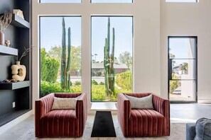 A modern living room with two red velvet armchairs, a black rug, and large windows with a view of cacti and greenery.