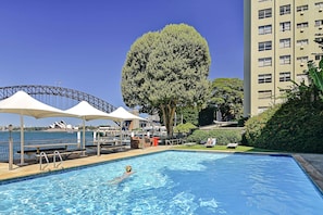 A swimming pool with a person swimming, a large tree, and a building with multiple windows.