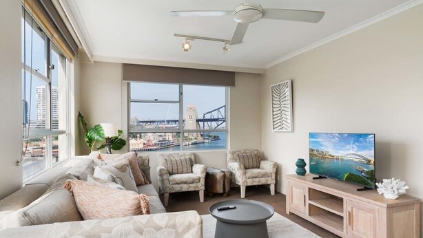 A modern living room with a sofa, armchairs, a coffee table, a TV on a wooden stand, and a large window with a view of a bridge and buildings. - Harbourside #53 (McMahons Point)