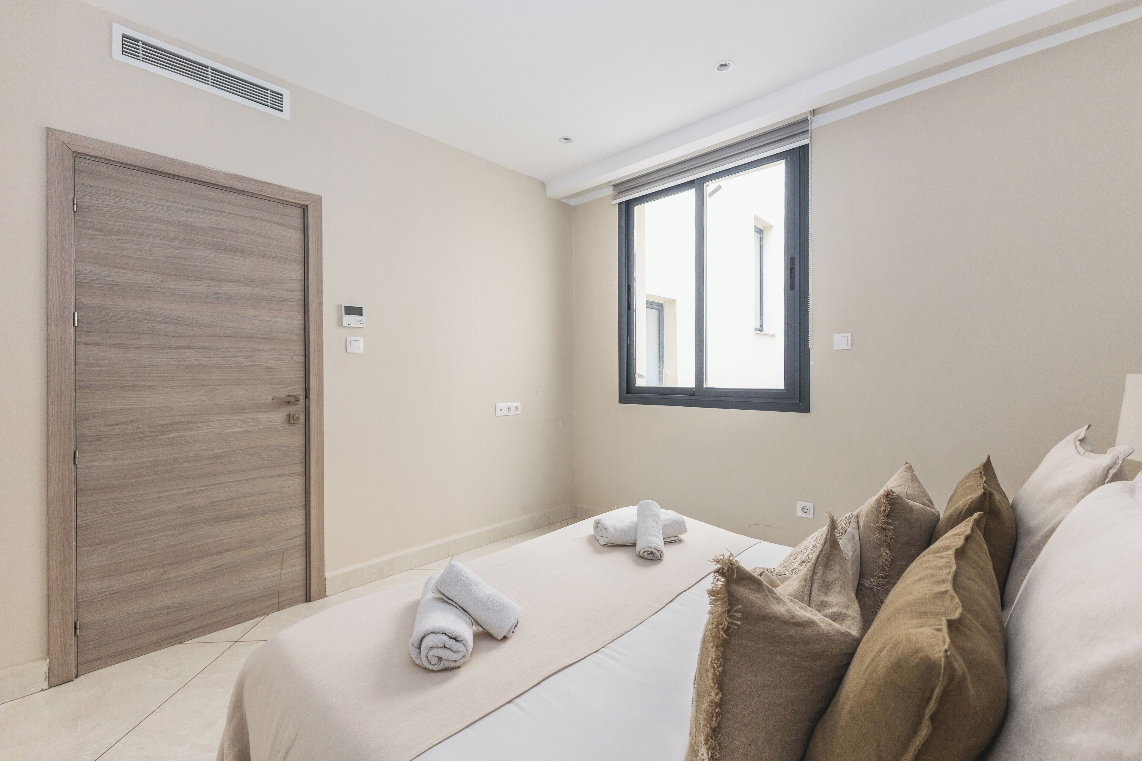 A hotel room with a large window, a bed with rolled towels, a wooden door, and a thermostat on the wall.