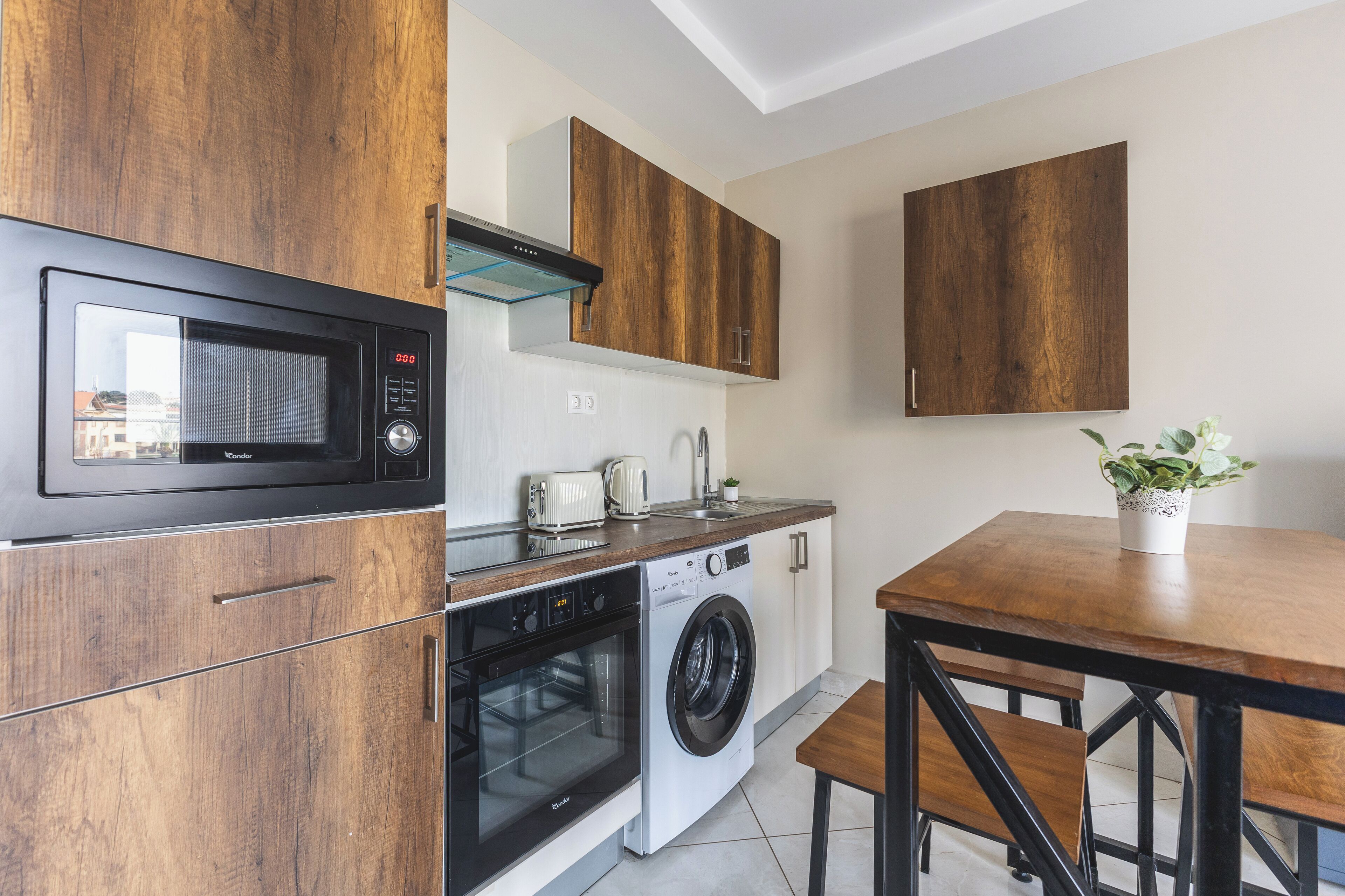 Eine kompakte Küche mit Mikrowelle, Backofen und Waschmaschine. Ein Holztisch und eine Bank bieten Platz, sowie ein Blumentopf auf dem Tisch.
