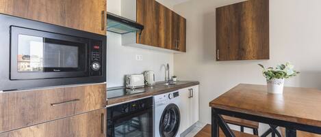 Eine kompakte Küche mit Mikrowelle, Backofen und Waschmaschine. Ein Holztisch und eine Bank bieten Platz, sowie ein Blumentopf auf dem Tisch.